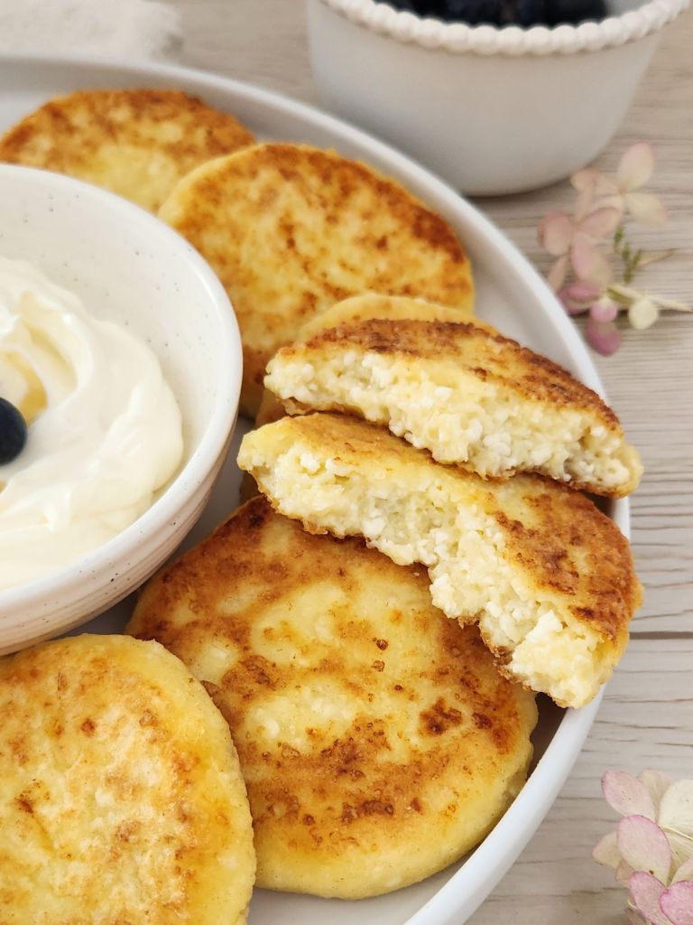 Golden cottage cheese pancakes served on a white plate with yougurt and blueberries with one pancake broken in half to see the texture