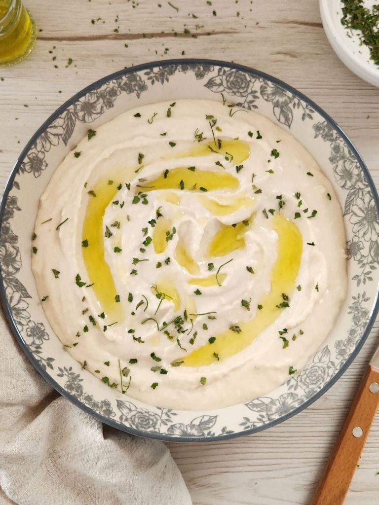 Bowl of creamy white bean hummus drizzled with olive oil and sprinkled with chopped parsley, served on a light wooden table with a linen napkin.