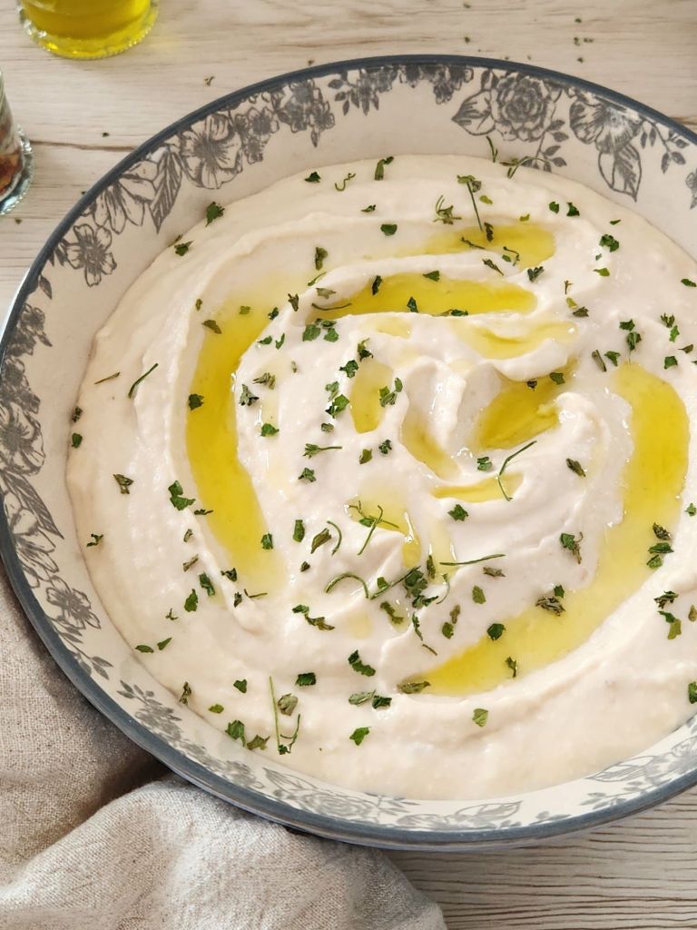 Bowl of creamy white bean hummus drizzled with olive oil and sprinkled with chopped parsley, served on a light wooden table with a linen napkin.