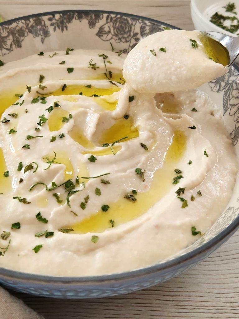 Close-up of creamy white bean hummus with a spoon showing its smooth, silky texture, drizzled with olive oil and topped with fresh parsley.