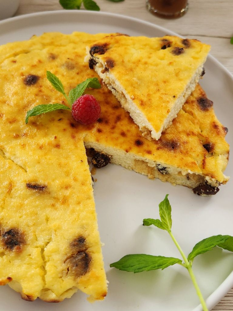 High-protein cottage cheese breakfast cake on a white plate, with a slice lifted to show soft, creamy texture, golden top, and raisins, garnished with fresh mint and a raspberry.