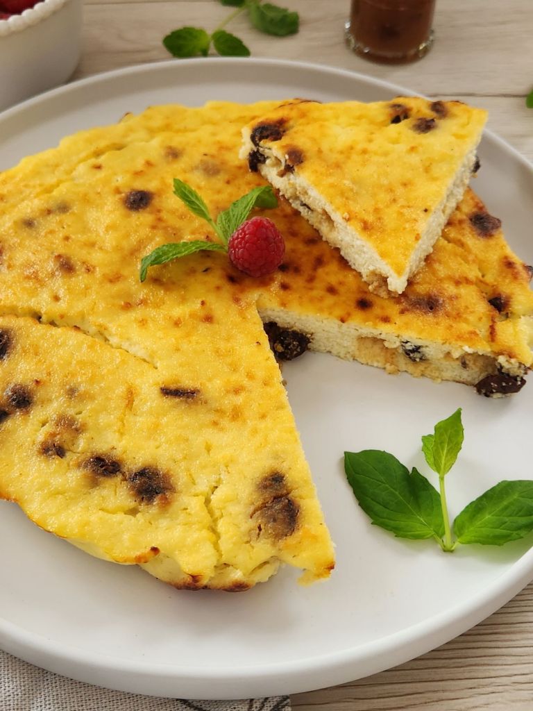 High-protein cottage cheese breakfast cake on a white plate, with a slice lifted to show soft, creamy texture, golden top, and raisins, garnished with fresh mint and a raspberry.