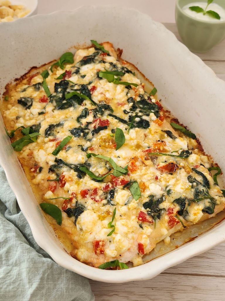 Baked salmon topped with feta cheese, sautéed spinach, parmesan and roasted bell peppers in a white baking dish.