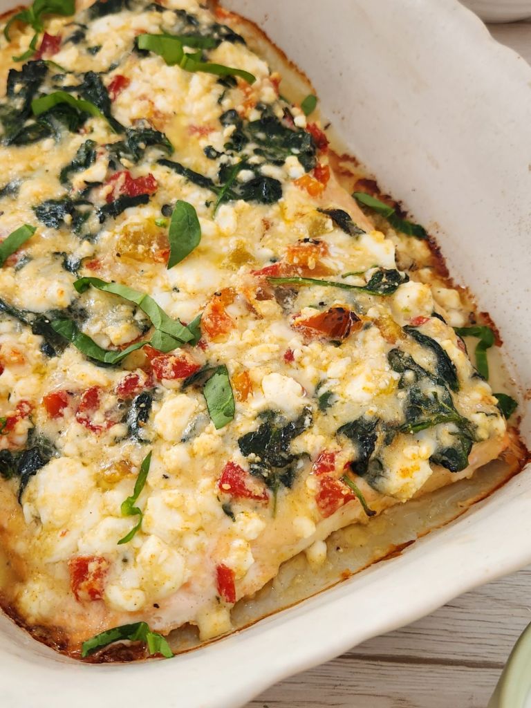 Close-up of baked salmon topped with feta cheese, sautéed spinach, parmesan and roasted bell peppers in a white baking dish.