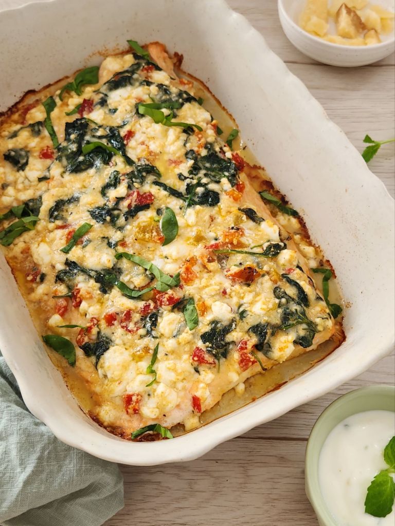 Baked salmon topped with feta cheese, sautéed spinach, parmesan and roasted bell peppers in a white baking dish.