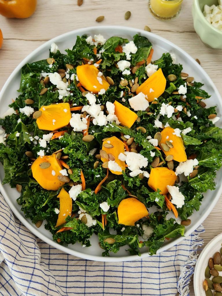 Kaki and kale salad with pumpkin seeds and feta on a white plate, topped with fresh orange dressing