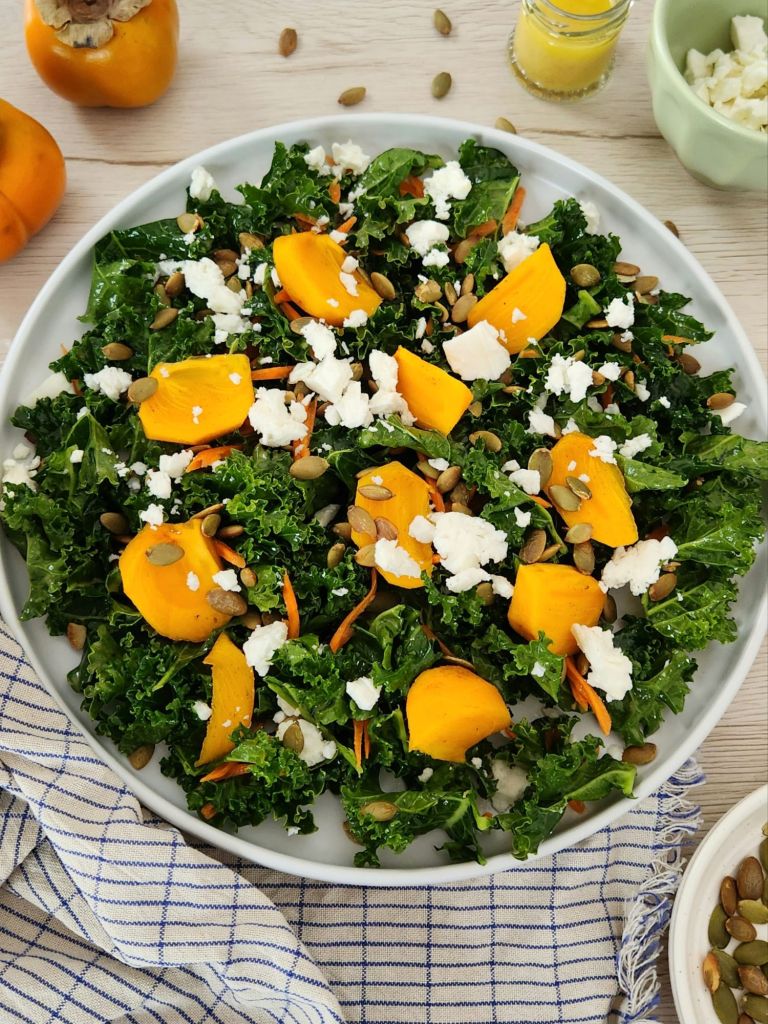 Kaki and kale salad with pumpkin seeds and feta on a white plate, topped with fresh orange dressing.