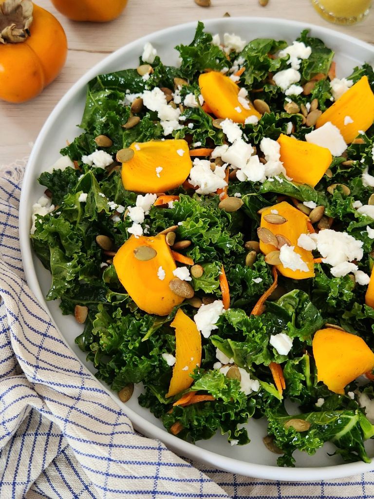 Close-up of kaki and kale salad with pumpkin seeds, feta, and citrus orange dressing showing fresh texture and vibrant colors.