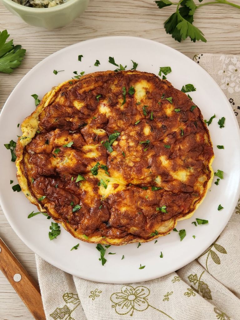 Homemade tomato and onion omelette made with eggs, milk, and grated cheese served on a white plate and topped with fresh parsley 