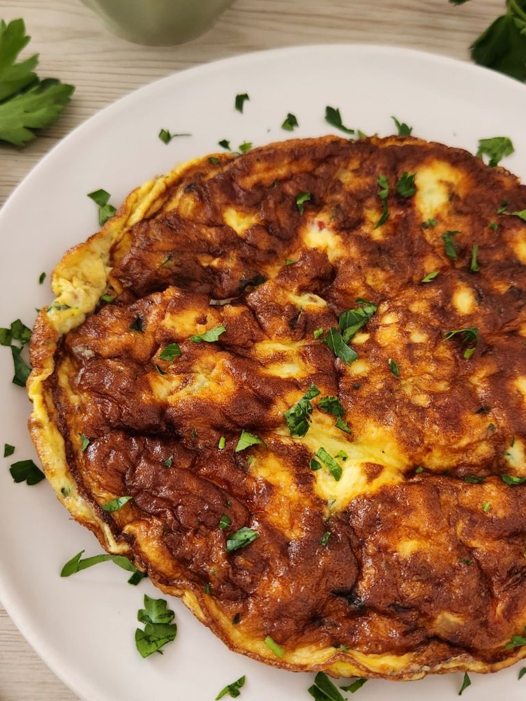 Homemade tomato and onion omelette made with eggs, milk, and grated cheese served on a white plate and topped with fresh parsley 