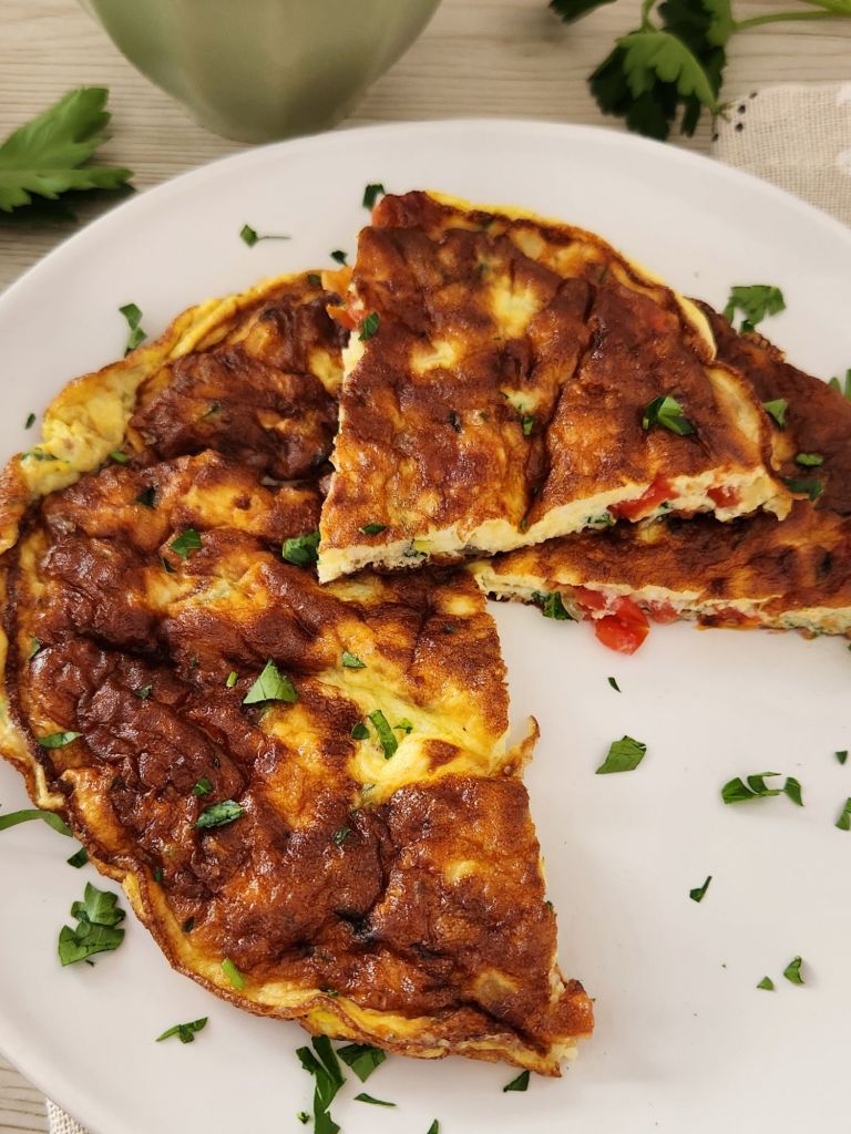 Homemade tomato and onion omelette with eggs, milk, and grated cheese, served on a white plate, garnished with parsley, with one piece cut