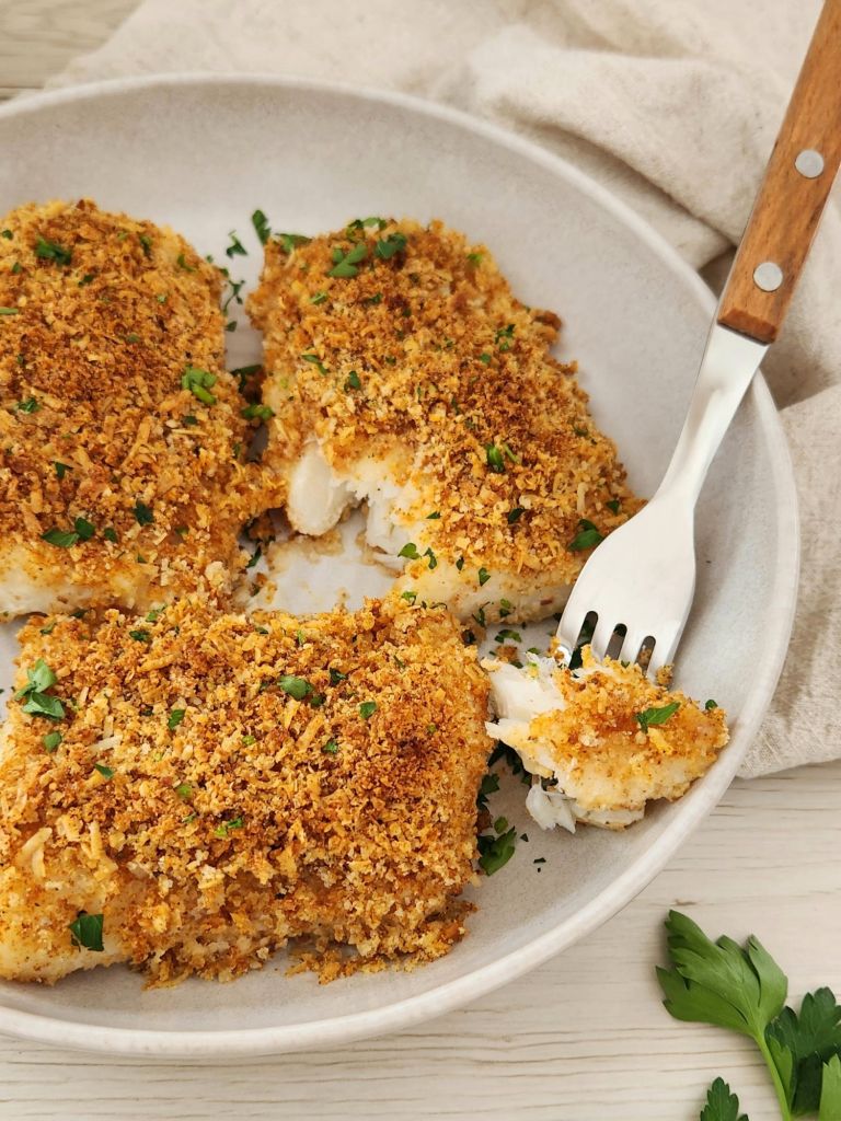 Crispy golden fillets of baked cod served on a beige plate and topped with parsley and one broken fillet to see texture