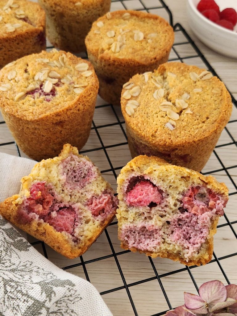 Gluten-free raspberry almond muffins on a cooling rack, with two muffins cut open to show the soft crumb and juicy raspberries inside.