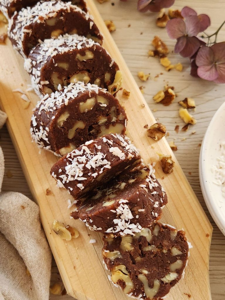 Slices of healthy chocolate salami with walnuts, covered in shredded coconut, served on a wooden board