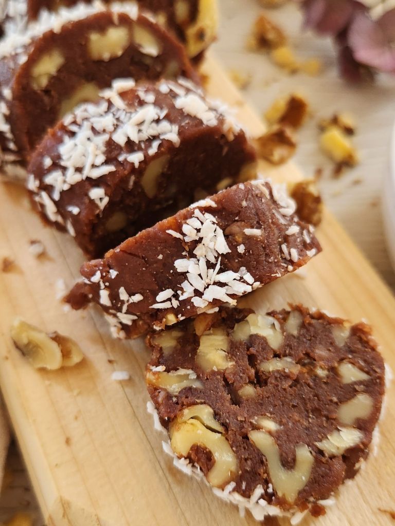 Slices of healthy chocolate salami with walnuts, covered in shredded coconut, served on a wooden board