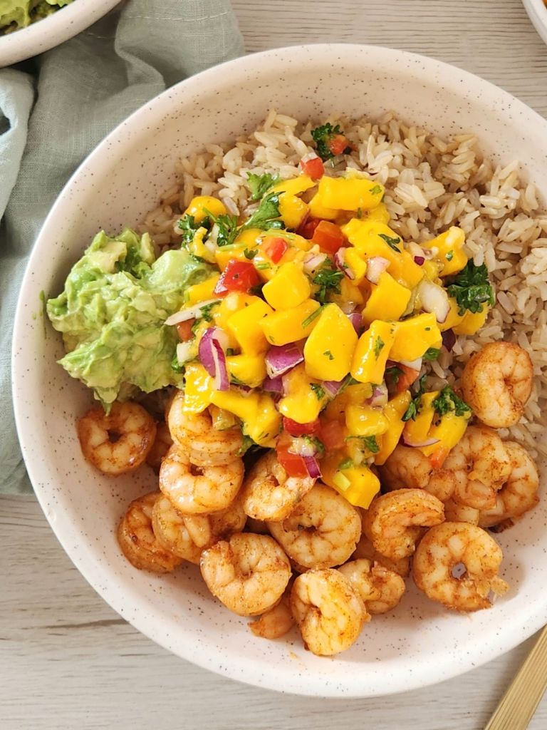 Brown cooked rice with cooked shrimp, mashed avocado and mango salsa served in a beige bowl