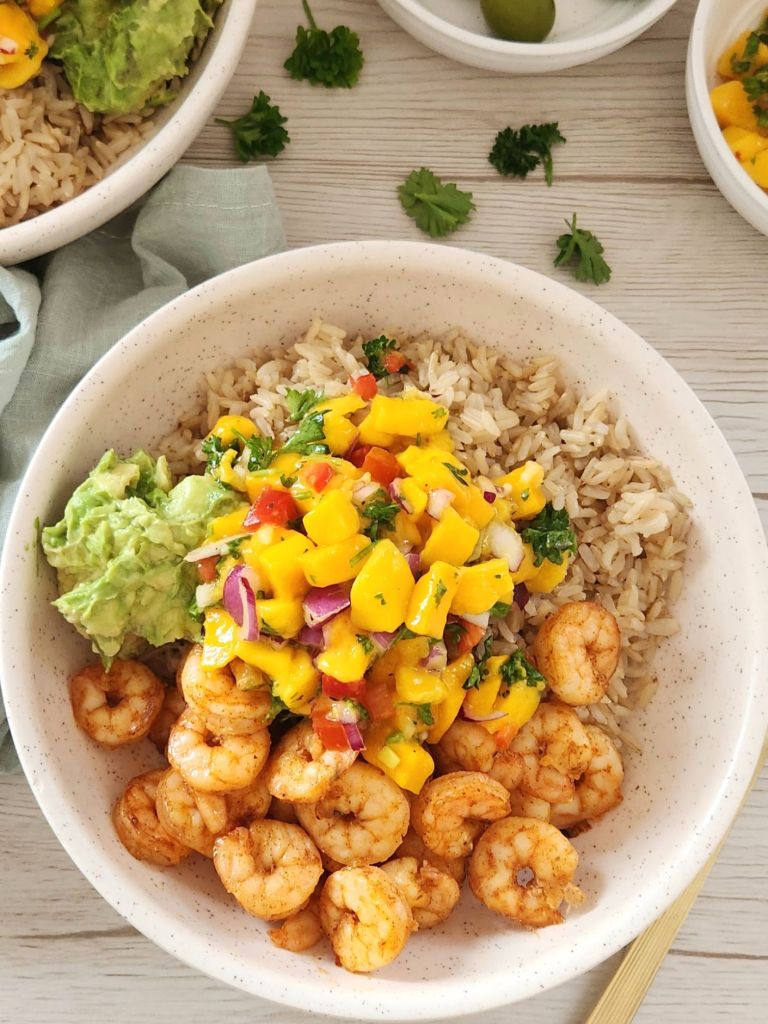 Brown cooked rice with cooked shrimp, mashed avocado and mango salsa served in a beige bowl