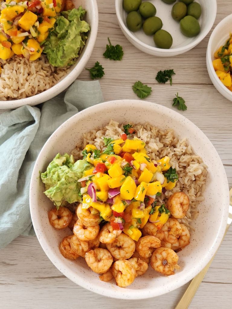 Brown cooked rice with cooked shrimp, mashed avocado and mango salsa served in a beige bowl
