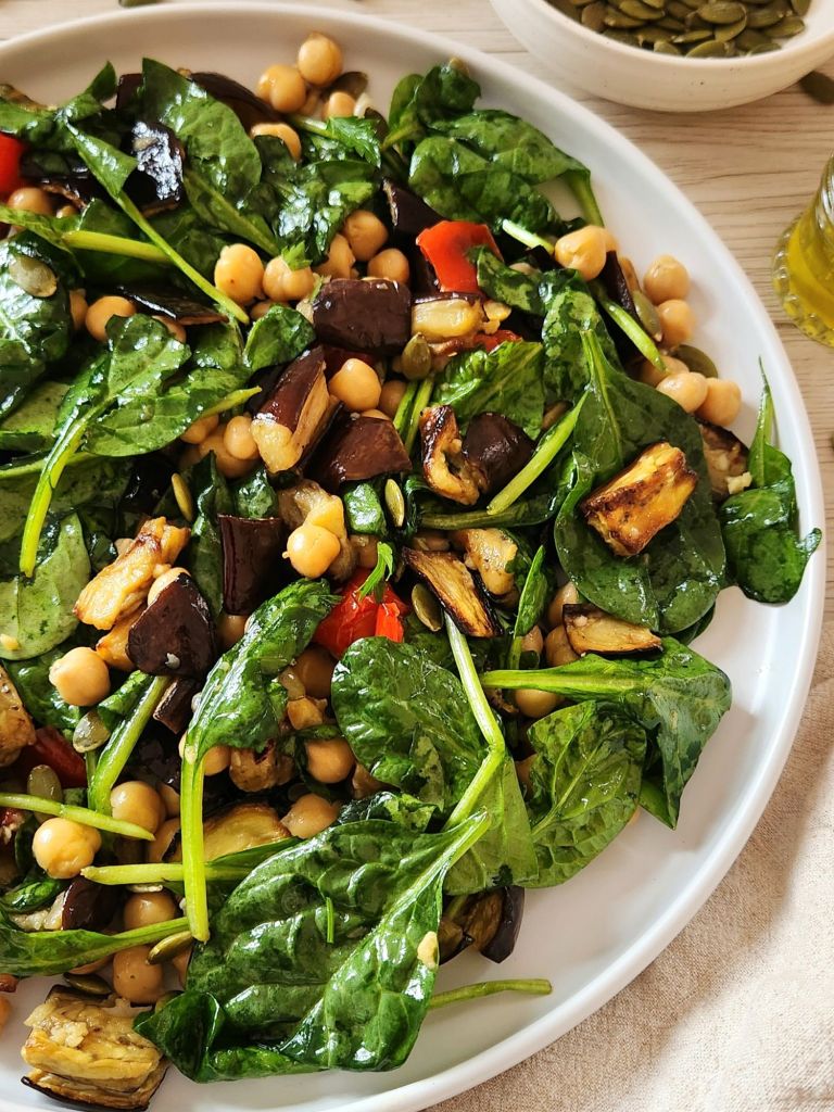 warm salad with roasted eggplant with chickpeas, spinach, red pepper, pumpkin seeds served on a white plate
