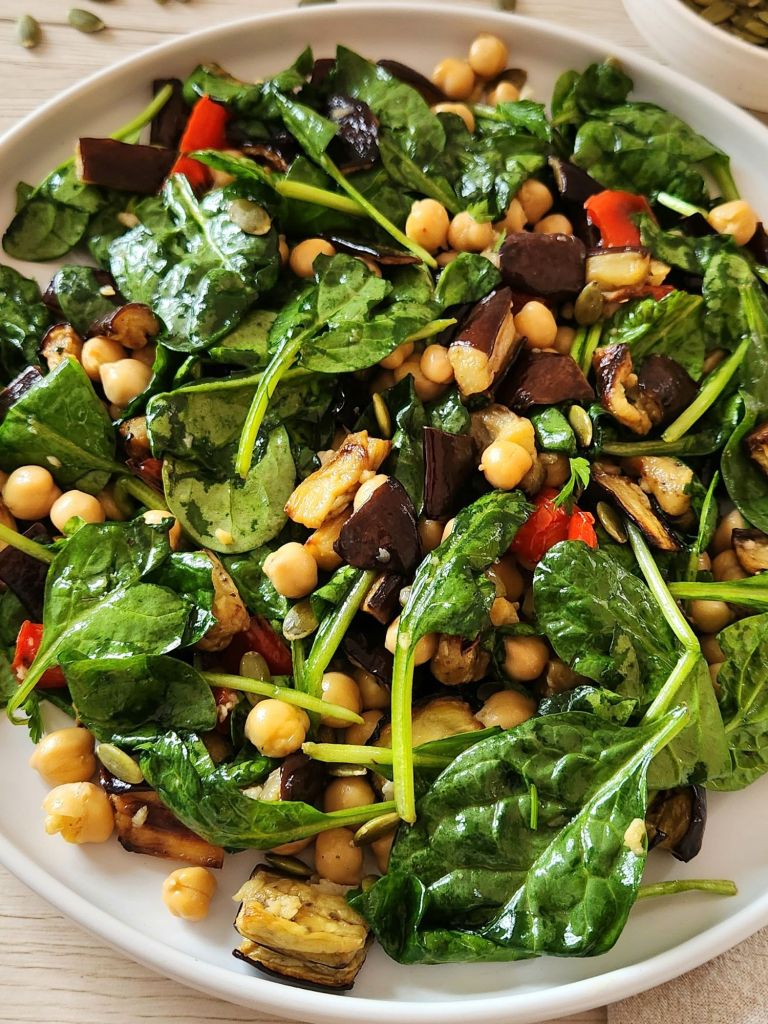 warm salad with roasted eggplant with chickpeas, spinach, red pepper, pumpkin seeds served on a white plate