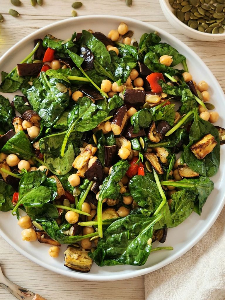 warm salad with roasted eggplant with chickpeas, spinach, red pepper, pumpkin seeds served on a white plate