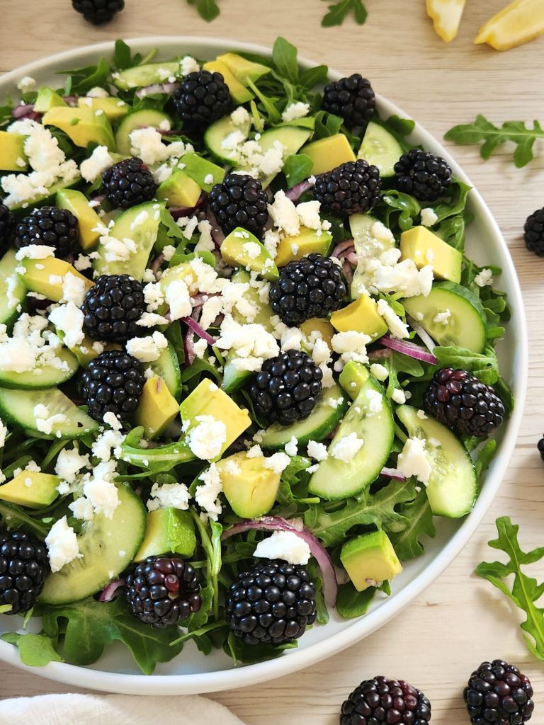 Fresh salad with blackberries, arugula, avocado, cucumber slices and crumbled feta served in a white plate