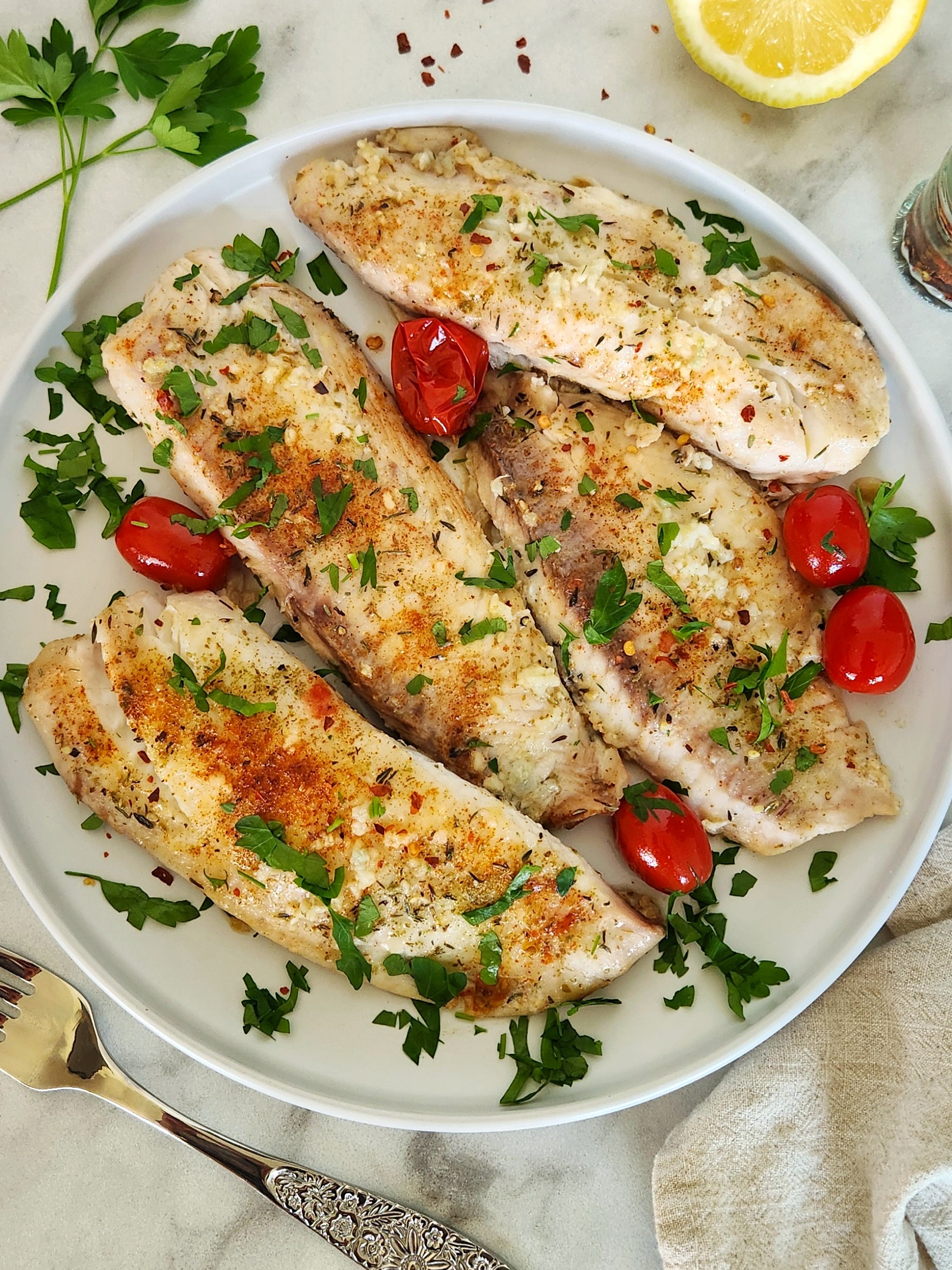 Healthy baked tilapia topped with garlic, herbs, and lemon on a white plate with cherry tomatoes and fresh parsley