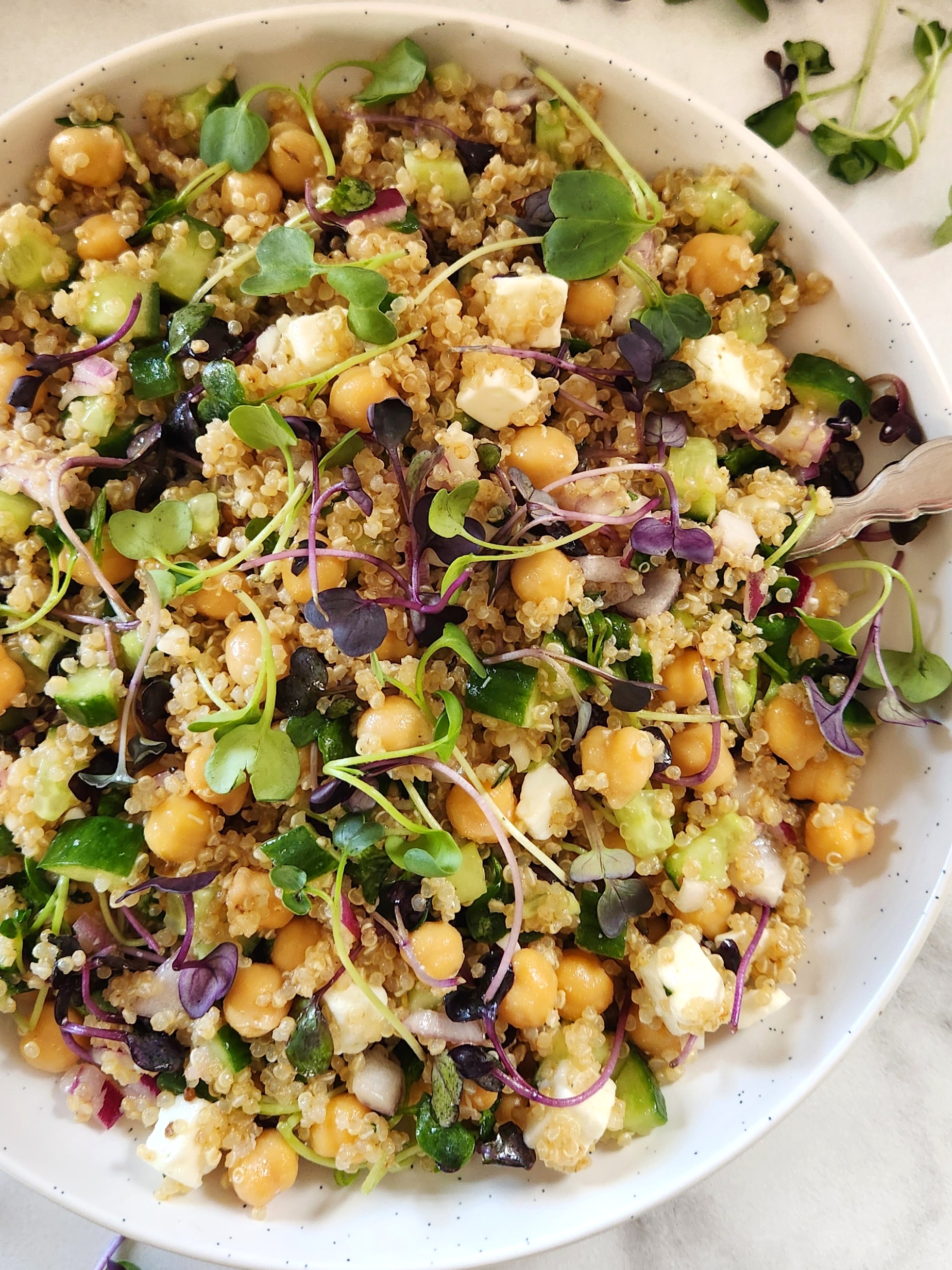 Healthy quinoa chickpea feta salad with cucumber, red onion, fresh microgreens, and a lemon olive oil dressing served in a white bowl.