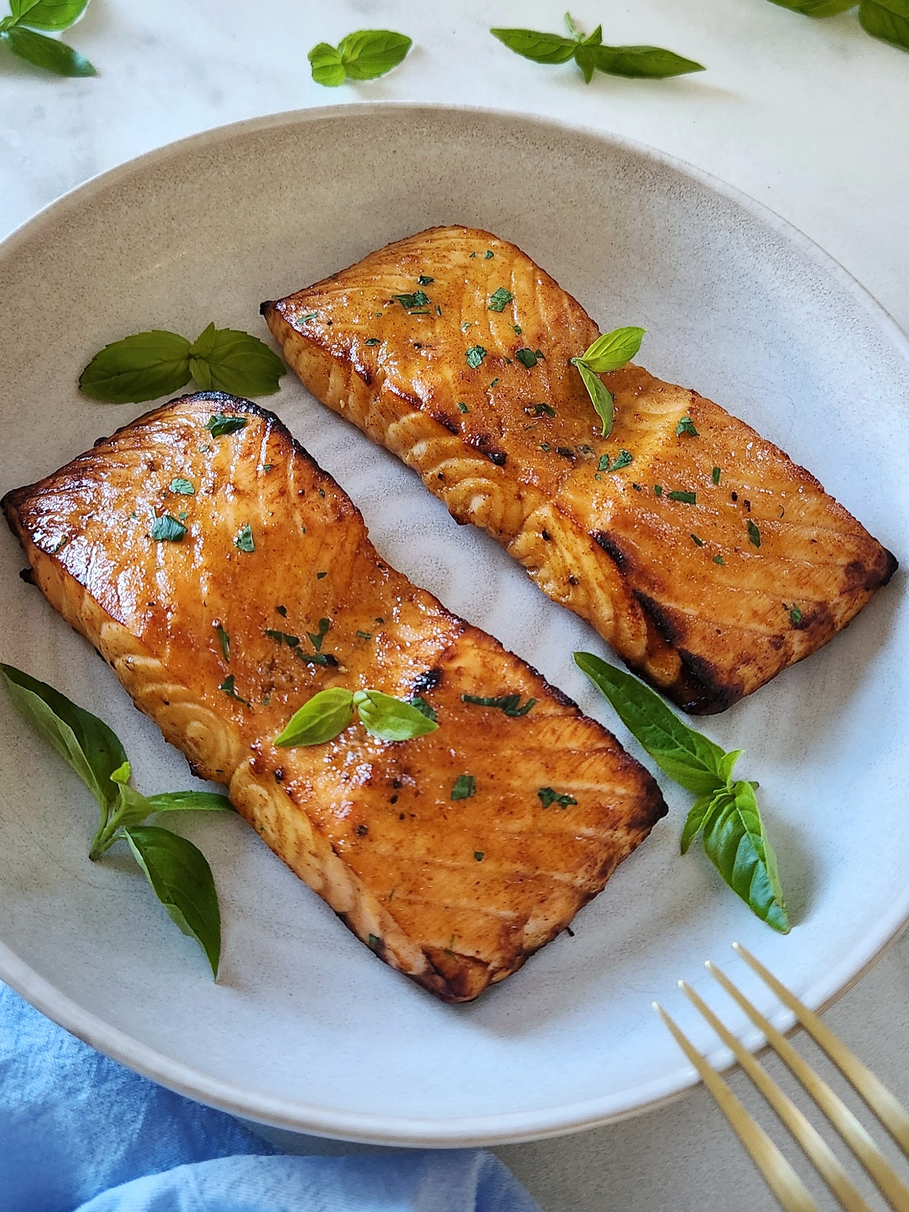 Two golden-brown air fryer salmon fillet garnished with fresh basil on a beige plate.